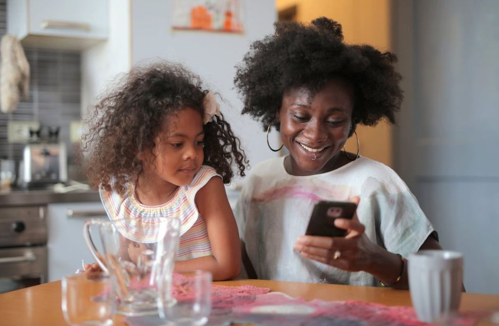 Woman sitting at a kitchen table with her daughter in her lap while using a mobile phone