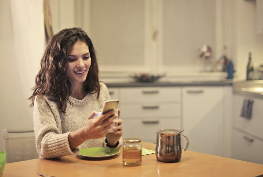 woman sitting a kitchen table drinking tea and browsing on her mobile phone