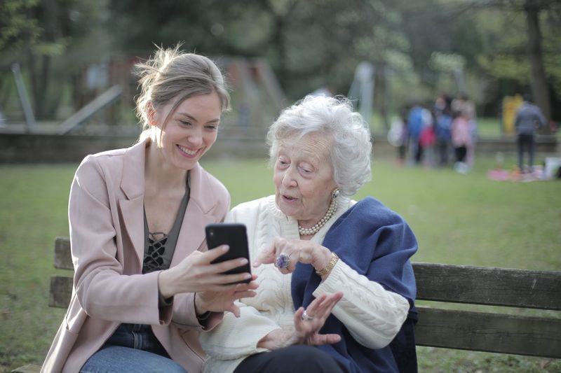 Young woman smiling while showing something on her phone to an elderly woman sitting next to her on a park bench
